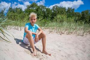 Model posing Handsome teenager boy of European appearance with blond hair in white shorts, and a blue T-shirt sits on a beach, and looks thoughtfully into the distance. Summer vacation concept.Summer travel concept.Copy space. photo