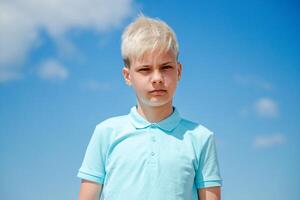 Portrait handsome teenager boy of European appearance with blond hair in a blue T-shirt stands behind blue sky, and looks to the camera. Summer vacation concept.Handsome teenager boy portrait concept.Copy space. photo