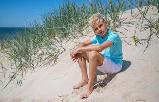 Handsome teenager boy of European appearance with blond hair in white shorts, and a blue T-shirt sits on a beach, and looks to the camera. Summer vacation concept.Summer travel concept.Copy space. photo