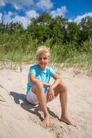 Handsome teenager boy of European appearance with blond hair in white shorts, and a blue T-shirt sits on a beach, and looks thoughtfully into the distance. Summer vocation concept.Summer travel sale concept.Vertical photo. photo