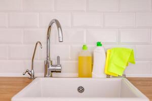 White plates, glass cups and forks in a clean white sink and two bottles of detergent and microfiber rags on a wooden table in a white kitchen photo