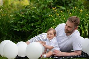 un papá y su pequeño hijo mentira en el césped rodeado por blanco globos del padre día. foto