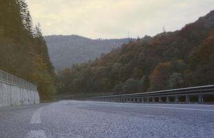 Road in autumn forest at sunset in Carpathian mountains, Ukraine. Beautiful mountain roadway with orange tress and high rocks. Landscape with empty highway through the woods in fall photo