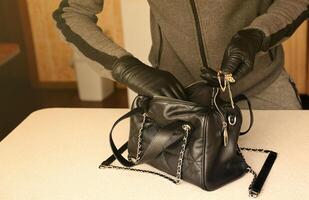 ladrón con traje negro y guantes que se ven en una bolsa de mujer robada abierta. el ladrón saca un collar de oro del bolso de una mujer en la cocina foto