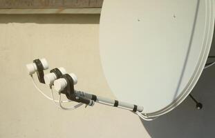 White satellite dish with three converters mounted on residental building rooftop concrete wall. Satellite television photo
