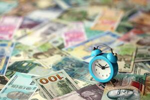 Small alarm clock on many banknotes of different currency. Background of time and money photo