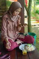 Side view of a Muslim woman eating fried rice while playing on her cellphone in a sitting position with her legs folded photo