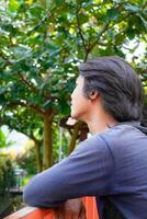 side view of a man sitting pensively with his hands resting on a security fence against a backdrop of trees. photo