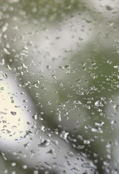 A photo of rain drops on the window glass with a blurred view of the blossoming green trees. Abstract image showing cloudy and rainy weather conditions
