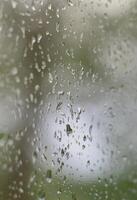 A photo of rain drops on the window glass with a blurred view of the blossoming green trees. Abstract image showing cloudy and rainy weather conditions