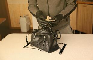Robber in black outfit and gloves see in opened stolen women bag. The thief takes out the gold and money from a womans handbag in kitchen photo