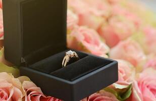 Gold diamond engagement ring in black box case among big amount of roses in big bouquet close up photo