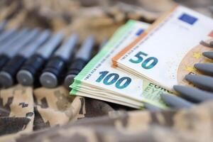 Bundles of euro bills and a machine gun belt on the camouflage uniform of a Ukrainian soldier. Concept of war financing, bribe or donation photo