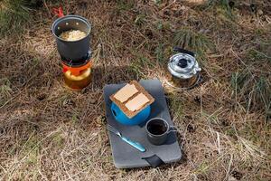 Tourist's breakfast in nature, camping food lying on the ground, butt seat, bowler with Chinese noodles, kettle with hot tea, cheese sandwiches, coffee mug, tourist dishes photo