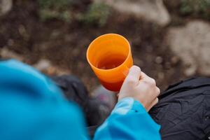 jarra en un mujer mano parte superior vista, acampar utensilios platos a un picnic, un niña bebidas café en naturaleza en un caminata, un naranja vaso hecho de polietileno, un eco plato articulo foto
