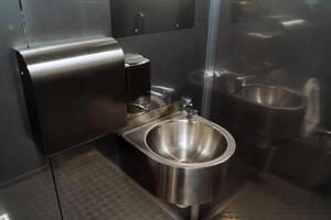 A metal washbasin hangs in a public toilet, an anti-vandal toilet on the street, metal walls of the room, a soap dispenser, a towel holder. photo