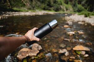 The guy holds a thermos in his hand against the background of the river, his hand holds a thermo bottle. trekking on a mountain river alone, hiking in extreme conditions. photo