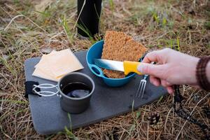 A tourist's hand holds a knife for slicing cheese, a man's breakfast on a hike in the woods, a cup of hot tea, a tourist lunch in nature, food in a camp, cheese slices. photo