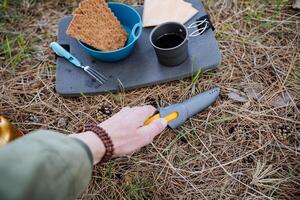 A tourist's hand pulls out a knife from a cover, breakfast in nature, camp food, crackers with cheese, tea in a glass, lunch in the camping forest, a knife in the hand of a bushcraft. photo