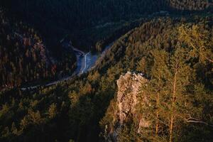 el ver desde el montaña a el rock iluminado por el sol, el río fluye abajo el montaña, el paisaje de el taiga bosque, pino árboles, Roca permanecer. foto