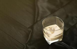 Glass of Irish cream baileys liqueur with ice cubes on dark fabric and wooden bar table background. Homemade luxury elite alcohol cocktail. Place for text photo