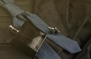 A bottle of mens cologne and cufflinks with blue tie lie on a black luxury fabric background on a wooden table. Mens accessories photo