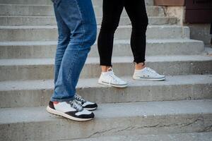 el pies de dos personas estar en el pasos de el escaleras, elegante nuevo zapatillas, moderno zapatos, casual estilo en ropa, desnudo tobillos, blanco zapatillas, azul vaqueros. foto