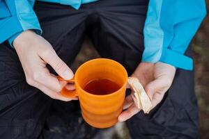 desayuno excursionismo emparedado con queso en mano sostener un jarra de té, bebida un caliente bebida en naturaleza, calentar arriba con calentar té, delicioso almuerzo a el cámping, naranja vaso foto