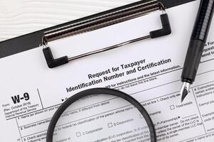 IRS Form W-9 Request for taxpayer identification number and certification blank on A4 tablet lies on office table with pen and magnifying glass photo