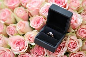 Gold diamond engagement ring in black box case among big amount of roses in big bouquet close up photo