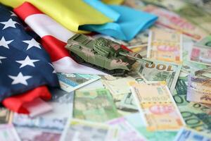 Toy tank on US flag and ukrainian flag on many banknotes of different currency photo