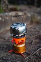 A metal kettle stands on a tourist burner in the forest, camping gas, tourist equipment, boiling water, hot tea, the handle is raised up. photo