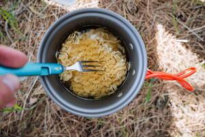 Delicious food on a hike, camping pot with hot meals, Chinese noodles cooked on a burner, trekking in the mountains, eating with a fork, a pot with a handle, tourist food, pasta in the water. photo