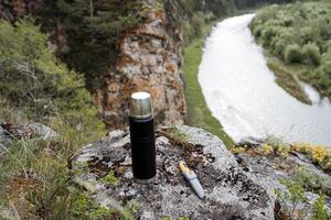 bebida té con un ver de el río, un termo soportes en un Roca alto en el montañas, un verano vacaciones en naturaleza, cámping en el borde de un acantilado, un cuchillo acostado en un roca, descanso en el bosque. foto