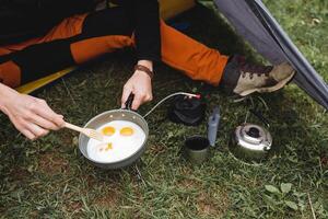 A tourist sits in a tent frying scrambled eggs in a frying pan, eating on a hike, cooking breakfast in the mountains, camping equipment utensils for climbing, stirring with a fork. photo