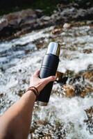 Hand holds a thermos with tea, camping equipment for hiking, mountain hiking, water tank, vacuum bottle, mountain stream, bracelet on the arm photo