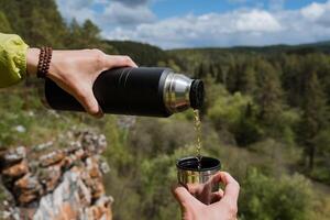 bebida té en un caminata en contra el antecedentes de el montañas, manos participación un termo con un taza, verter un caliente bebida desde un termo botella dentro un taza, excursionismo en el bosque, un viajeros desayuno. foto