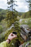 A man's hand holds a glass of coffee against the background of the forest, a rudraksha bracelet on the wrist, a hipster drinks tea on a hike, a thermo glass, lift a cup up photo