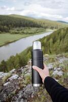 A person's hand holds a metal thermos with tea against the background of nature, a tourist shows a bottle with a hot drink, a black steel thermos. photo