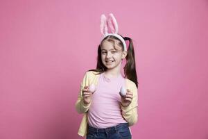 Young toddler smiling and wearing bunny ears in front of camera, presenting her handmade painted pink eggs for easter holiday tradition. Cheerful cute girl in studio shows ornaments. photo