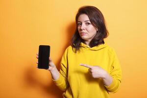 Brunette lady showing cellphone with empty screen at camera in studio. Woman pointing at mobile phone with isolated mockup template on display. Person standing over orange background photo