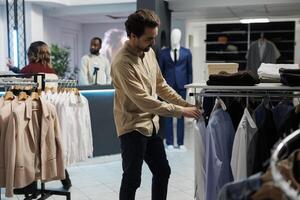 joven hombre cliente examinando vestir opciones en ropa Tienda Departamento mientras elegir formal tener puesto. comprador comprobación camisas mientras explorador estante en compras centro comercial sala de exposición foto
