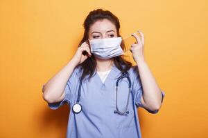 enfermero facultativo poniendo en cara máscara para proteccion en frente de cámara. hembra médico médico vistiendo matorrales y estetoscopio teniendo la seguridad máscara en contra coronavirus durante pandemia. foto