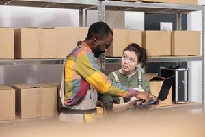 Multi ethnic stockroom supervisor packing parcels and managing delivery operations. Colleagues working in post office storage, standing beside cardboard boxes shelves preparing orders in storage room photo