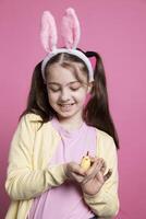 Small young kid showing a cute stuffed chick toy in studio, joyful little girl with golden arrangement over pink background. Youngster with sweet fluffy ears for easter festivity in front of camera. photo