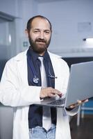 Portrait shot of a male physician in a white lab coat holding a digital laptop. Looking at camera is a caucasian doctor grasping a wireless computer in a hospital office room. photo