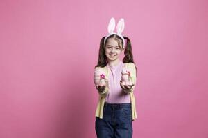 festivo dulce niña participación Pascua de Resurrección decoraciones y mullido juguetes en estudio, niño posando con un rosado huevo y un Conejo. adorable joven vistiendo coletas y conejito orejas terminado rosado antecedentes. foto