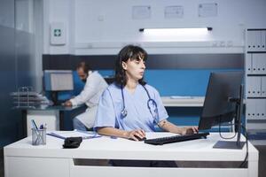 cucasian facultativo es mecanografía un médico reporte a un escritorio en un hospital oficina tarde a noche. hembra enfermero preparando médico cuidado mientras evaluando paciente enfermedad síntomas. foto