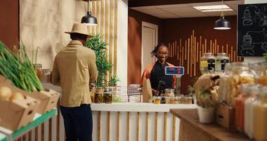 African american seller working at supermarket cash register counter, weighting organic additives free fruits and vegetables. Vendor selling fresh bio produce to male customer at checkout. Camera 2. photo