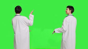 Physician pointing to the left or right to create web commercial standing against greenscreen backdrop, doing a marketing advertisement in studio. Woman medic in white coat shows an ad. Camera B. photo